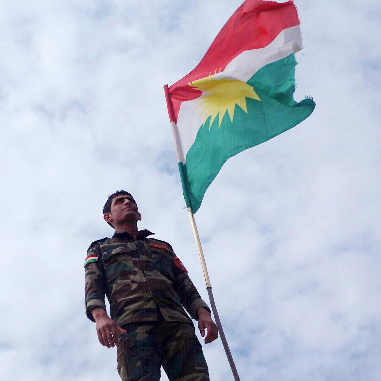در تصویر، یک سرباز در کنار پرچم کردستان ایستاده است. او لباس نظامی به تن دارد و پرچم با رنگ‌های قرمز، سفید، سبز و دایره زرد رنگ را که نماد هویت کردی است، به اهتزار درآورده است. پس‌زمینه آسمان ابری است و این ترکیب به تصویر حس قوی و نمادین می‌دهد.