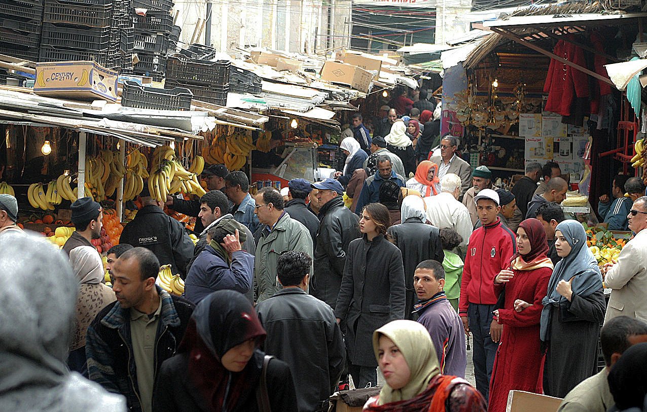L'image montre un marché animé, probablement dans une ville du Moyen-Orient ou d'Afrique du Nord. Des stands sont remplis de fruits, notamment des bananes et des oranges. On peut voir de nombreuses personnes, vêtues de vêtements variés, se déplacer entre les étals. L'atmosphère est animée, avec des groupes discutant et des clients choisissant des marchandises. Des éclairages pendent du plafond, ajoutant une ambiance particulière au lieu.