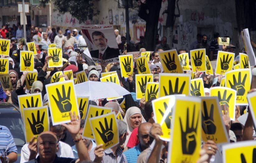 L'image montre une grande foule de personnes participant à une manifestation. Elles tiennent des affiches jaunes avec un symbole de main noire en faisant le signe qui pourrait représenter un mouvement ou une cause spécifique. La foule semble déterminée et engagée, et l'atmosphère est chargée d'énergie. On peut voir des pancartes et des slogans en arrière-plan. L'événement semble vouloir exprimer une opinion ou revendiquer des droits.