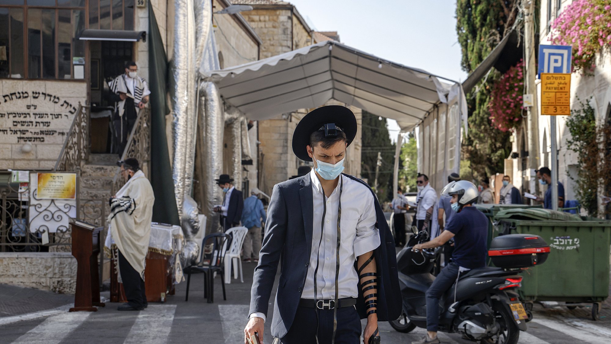 The image shows a street scene in an urban area, likely in a Jewish neighborhood. A young man wearing a mask and traditional attire, including a black hat and a suit, is walking while wearing tefillin (phylacteries) on his arm. In the background, there is a large tent set up for what appears to be a religious gathering, with several other individuals, some dressed in traditional clothing, visible. The setting is sunny, and there are various buildings, including signs and greenery, contributing to the atmosphere of the scene.