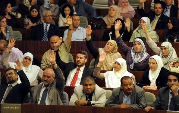 L'image montre un rassemblement dans un espace législatif, probablement au sein d'une assemblée ou d'un parlement. De nombreuses personnes sont présentes, avec une majorité levant la main, ce qui indique probablement un vote ou une approbation. Les participants semblent porter des tenues formelles, et certaines femmes portent le hijab. On observe une diversité de participants, tant en termes de genre que d'âge. L'environnement évoque une atmosphère de discussion et de prise de décision collective.