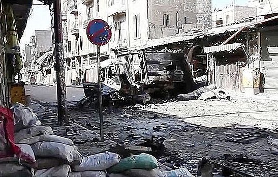 L'image montre une rue dévastée, probablement dans une zone de conflit. On peut voir des bâtiments endommagés et détruits, avec des débris éparpillés sur le sol. Des sacs de sable sont accumulés sur le côté, ce qui suggère des efforts de protection. Un panneau de signalisation est visible, mais l'environnement semble plutôt déserté et en ruines. L'atmosphère dégage un sentiment de désolation et de destruction.