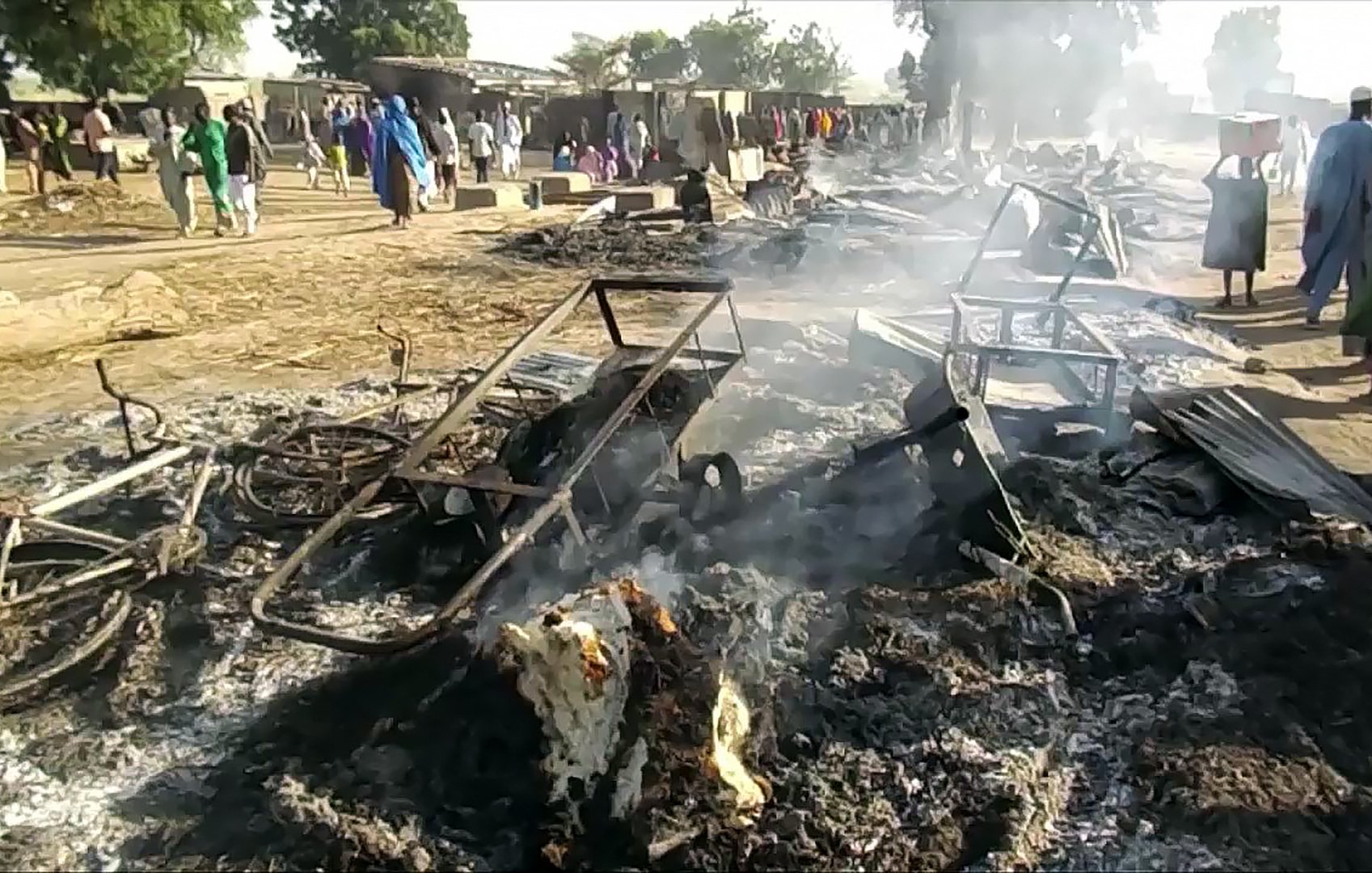 The image depicts a scene of destruction and aftermath, showing charred remains of structures and debris on the ground, with smoke rising. Various burned-out materials, such as metal frames and remnants of possibly furniture, are scattered across the area. In the background, a group of people can be seen, moving in what appears to be a community area, amidst the devastation. The setting suggests a recent fire or significant damage event, with the landscape appearing ashen and scattered with remnants of what once existed.