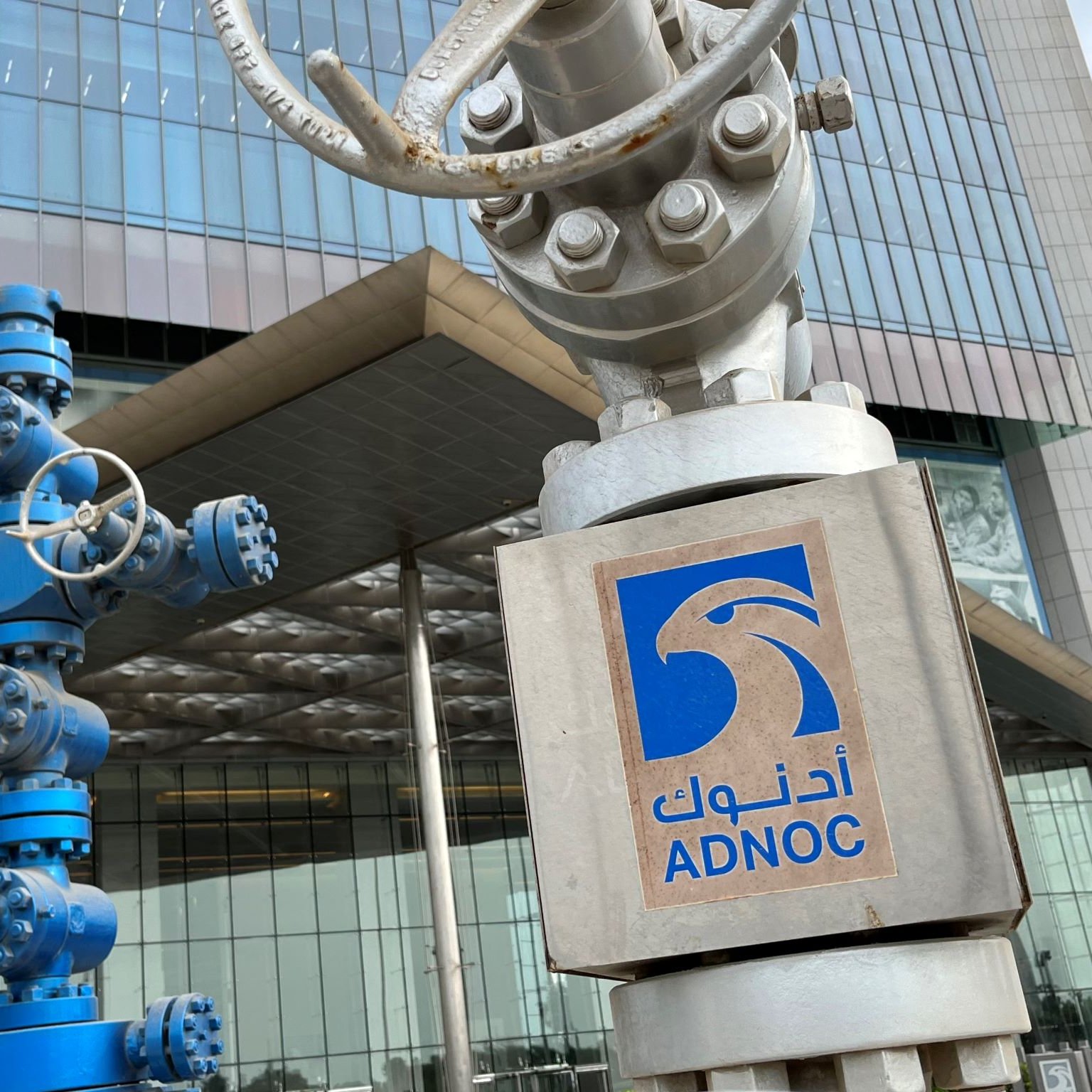 تصویر نشان‌دهنده نمای ساختمان شرکت نفت ابوظبی (ADNOC) است. در جلو، دو ساختار فلزی وجود دارد که شبیه به تجهیزات صنعتی و دکل‌های نفتی هستند. یکی از این تجهیزات به رنگ آبی و دیگری نقره‌ای است. در سمت راست تصویر، لوگوی ADNOC با نوشته عربی و انگلیسی قابل مشاهده است. ساختمان دارای ظاهری مدرن و شیشه‌ای است که عناصر طراحی معاصر را به نمایش می‌گذارد.