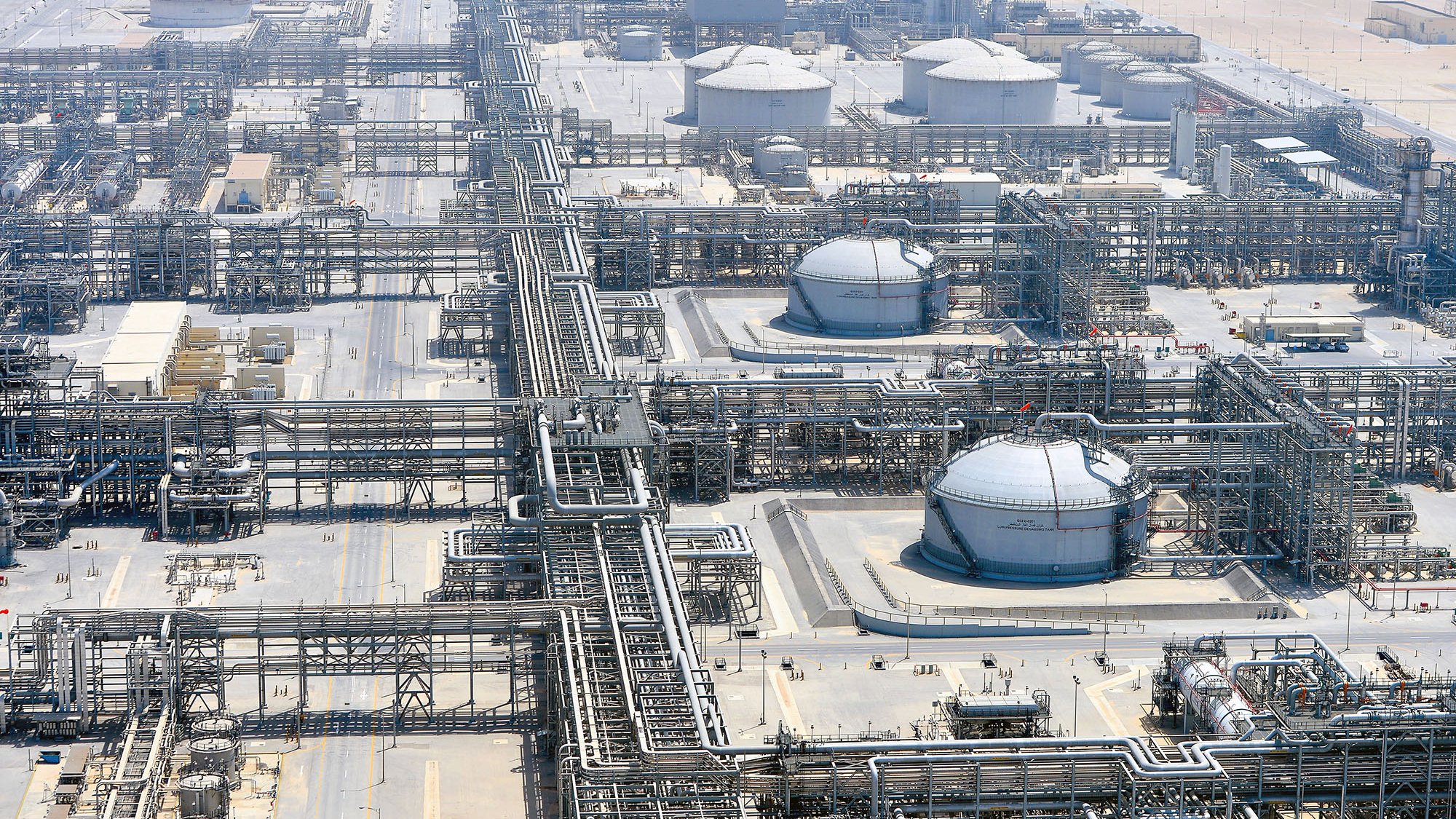 La imagen muestra una vista aérea de una planta industrial, probablemente relacionada con la producción o el refinado de petróleo. Se pueden observar grandes tanques de almacenamiento, tuberías extensas que se entrelazan por todo el área y estructuras de soporte. El entorno es desértico, con un paisaje árido al fondo, lo que sugiere que esta instalación podría estar ubicada en una región donde la extracción de recursos naturales es común. La organización y complejidad de las tuberías y tanques reflejan la sofisticación de la infraestructura industrial.