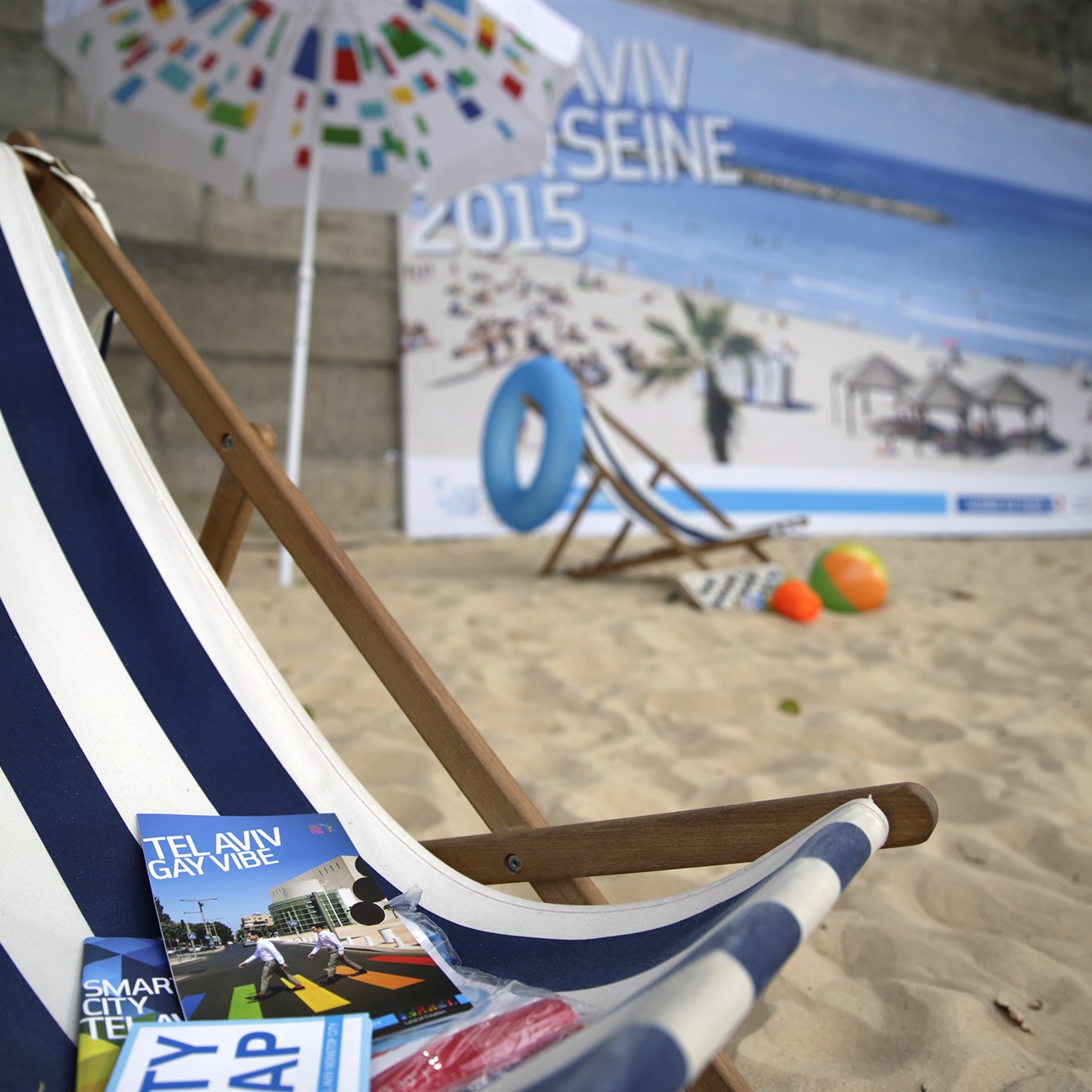 The image features a beach scene with a wooden deck chair adorned with blue and white stripes, set against a backdrop of a sandy area. In front of the chair are brochures for Tel Aviv, including one titled "Tel Aviv Gay Vibe" and another labeled "Smart City Map." The background includes a large poster depicting a sunny beach with umbrellas and people relaxing, suggesting a vibrant, beachside atmosphere.