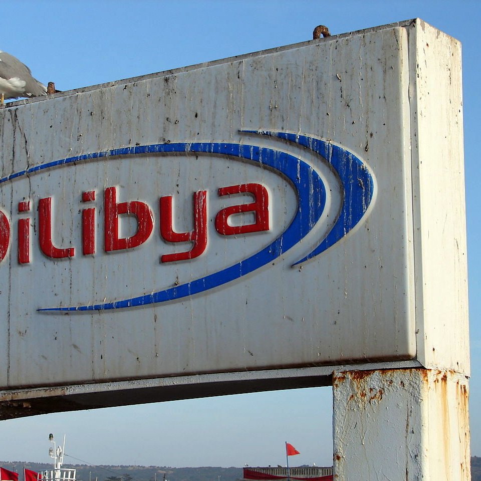 L'image montre un panneau publicitaire corrodé et usé portant le nom "Oil Libya" en lettres bleues et rouges. Au sommet du panneau, un goéland est posé. En arrière-plan, le ciel est clair, suggérant une belle journée. Le panneau semble être situé près de l'eau, probablement dans un port ou une zone côtière.