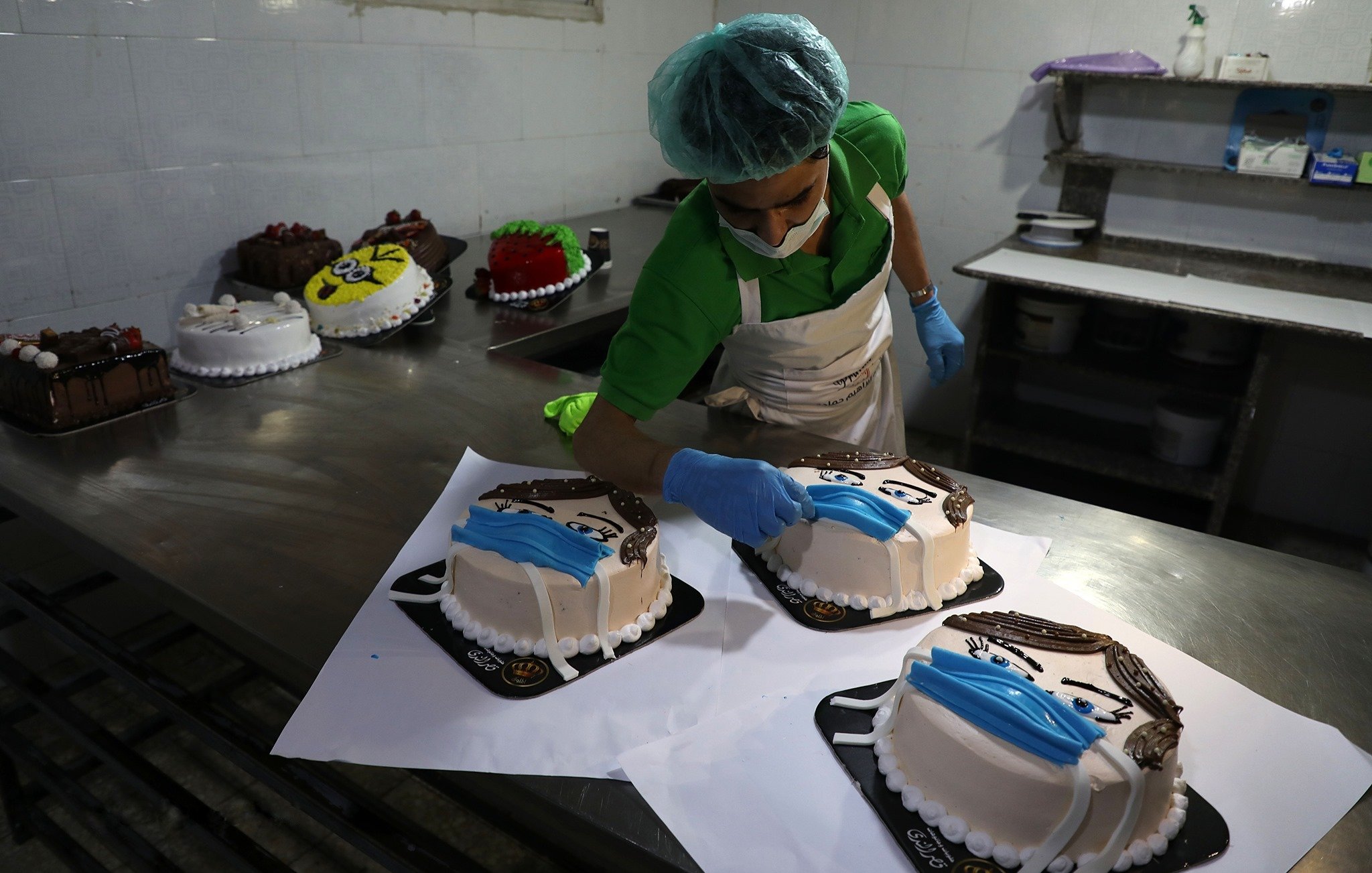 L'image montre un pâtissier en train de décorer des gâteaux dans une cuisine professionnelle. Il porte un bonnet de protection et des gants en latex. Sur la table, plusieurs gâteaux sont disposés, chacun ayant une décoration distincte, notamment des motifs qui ressemblent à des visages portant des masques. L'ambiance est soignée et les gâteaux sont présentés sur des plateaux noirs.