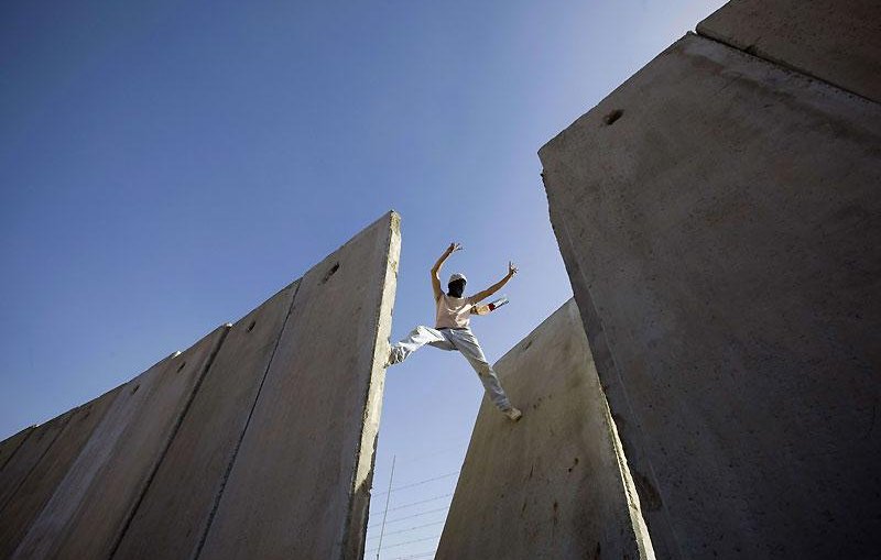 L'image montre une personne qui effectue une sorte d'acrobatie ou de saut entre deux grandes structures en béton. Le ciel est bleu, ce qui donne une impression d'ampleur et de liberté. La personne semble en mouvement, exploitant l'espace entre les murs. Cette scène peut évoquer des thèmes tels que le dépassement des obstacles ou la recherche de la liberté.