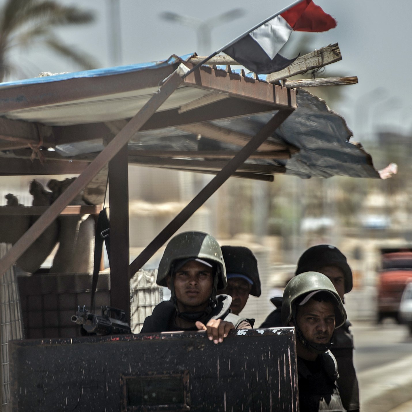 La imagen muestra a un grupo de soldados en un vehículo militar o en un puesto de control. Están vestidos con uniformes de combate y cascos, y algunos de ellos parecen estar armados. Al fondo, se pueden ver árboles y edificaciones, así como un cielo despejado. También hay un pequeño trozo de una bandera ondeando. La escena sugiere un ambiente de vigilancia o seguridad en una zona urbana.