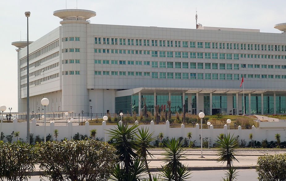 L'image montre un bâtiment moderne et imposant, probablement un centre administratif ou gouvernemental. Il est de couleur blanche avec de grandes fenêtres rectangulaires, donnant une impression de fonctionnalité et de sobriété. Le design architectural est contemporain, avec des formes géométriques claires. À l'avant, il y a un espace ouvert avec des éléments paysagers tels que des plantes et des lampadaires. On peut également voir un drapeau, ce qui pourrait indiquer qu'il s'agit d'un bâtiment officiel. L'environnement semble propre et bien entretenu.