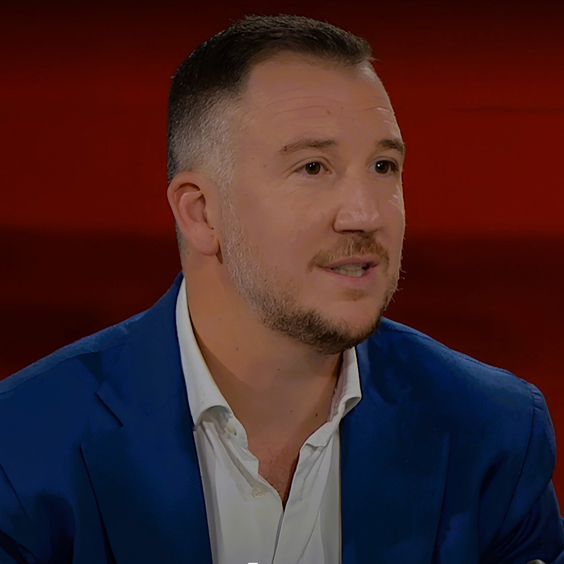 L'immagine mostra un uomo con capelli corti e una barba leggera, che indossa una giacca blu e una camicia bianca. Si trova di fronte a uno sfondo rosso. L'uomo appare in un contesto di colloquio o discussione, con un'espressione attenta e coinvolta.