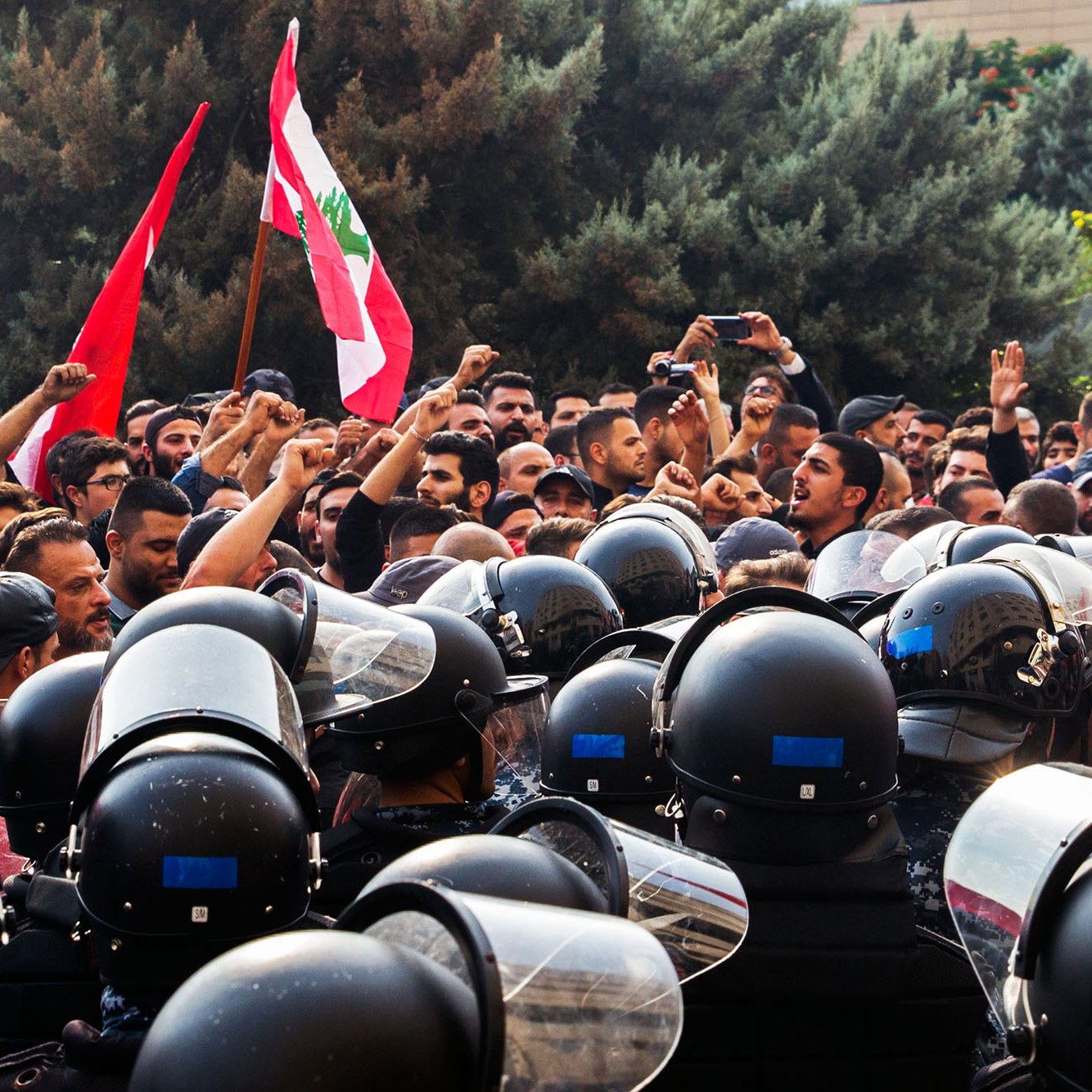 در این تصویر، یک تجمع اعتراضی در لبنان دیده می‌شود. گروهی از مردم در حال شعار دادن و به‌سوی دوربین نگاه می‌کنند، در حالی که پرچم‌های لبنان را در دست دارند. در مقابل آن‌ها، گروهی از نیروهای پلیس با لباس‌های ضدشورشی و کلاه‌خودهایی مشغول محافظت از جایگاه هستند. فضای تصویر تنش‌آلود و احساسات قوی در بین معترضین و نیروهای پلیس به وضوح حس می‌شود. در پس‌زمینه، ساختمان‌ها و درختان وجود دارند که به جو عمومی صحنه اشاره می‌کنند.