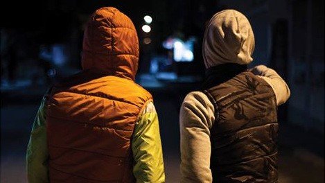 L'image montre deux personnes de dos, se tenant à proximité l'une de l'autre. L'une porte un gilet d'une couleur orange avec des manches vertes, tandis que l'autre est vêtu d'un gilet noir et d'une capuche grise. Ils semblent se trouver dans une rue sombre, avec quelques lumières au loin, ce qui donne une ambiance nocturne à la scène. L'une des personnes pointe quelque chose du doigt, suggérant une conversation ou une observation partagée.