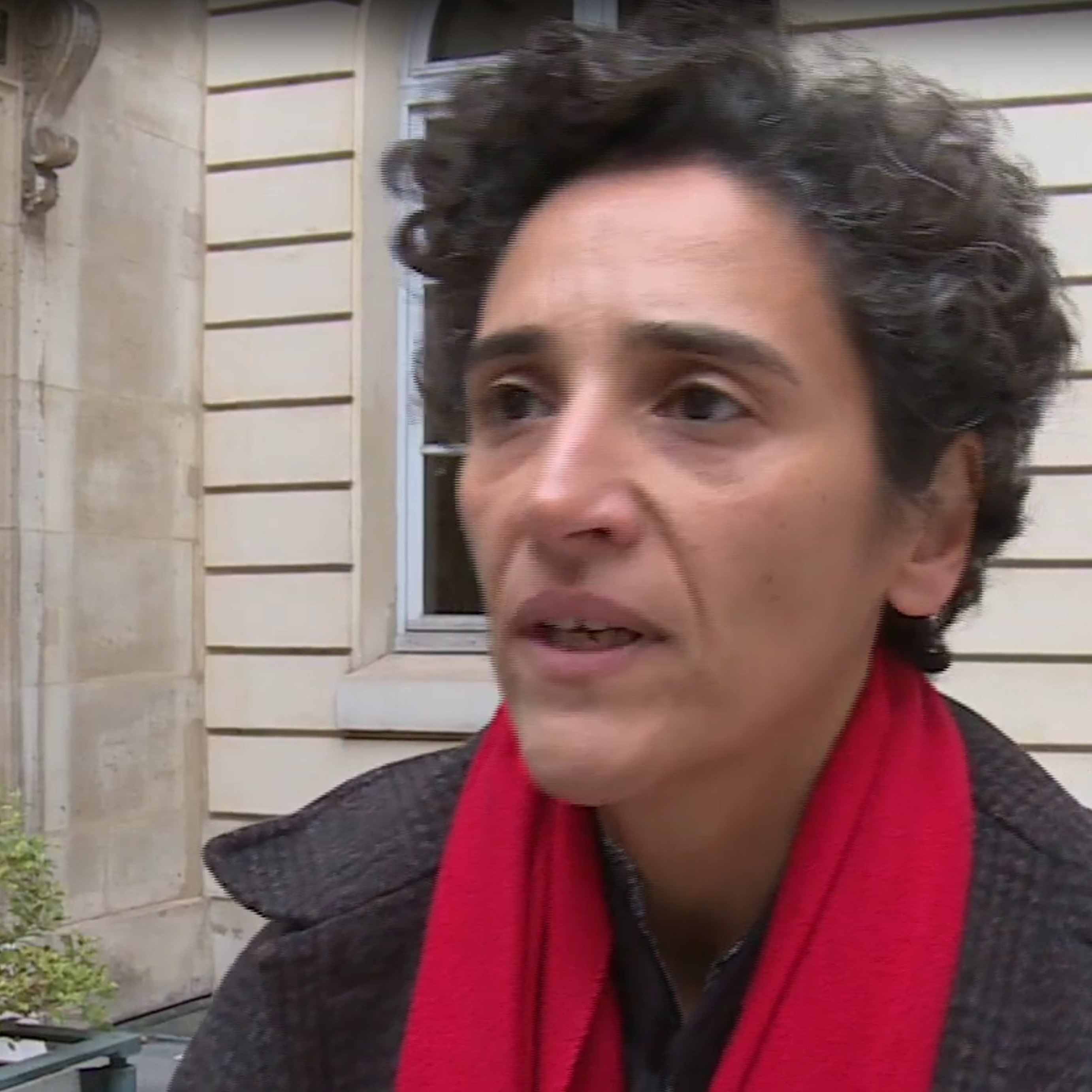 L'image montre une femme avec des cheveux bouclés, portant un manteau sombre et une écharpe rouge. Elle semble s'exprimer avec sérieux, peut-être dans le cadre d'une interview. En arrière-plan, on aperçoit un bâtiment avec des fenêtres et une porte d'entrée, ce qui donne une impression urbaine et architecturale.