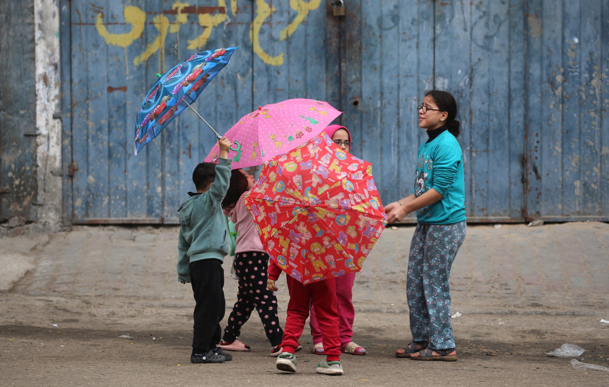 L'image montre un groupe d'enfants jouant dans la rue. Ils ont des parapluies colorés, certains avec des motifs amusants. Les enfants semblent joyeux et engagés dans leur jeu. En arrière-plan, il y a un mur bleu, qui donne une ambiance urbaine à la scène. Ils portent des vêtements confortables, adaptés à une activité en extérieur. L'atmosphère générale est légère et pleine de vie.