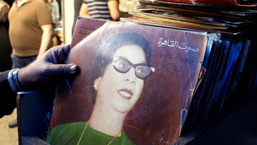 L'image montre une personne tenant un disque vinyle dans un marché. Le disque est de couleur foncée, avec une illustration d'une femme portant des lunettes et des vêtements vintage. En arrière-plan, on aperçoit d'autres personnes dans un environnement animé, probablement un marché ou une braderie, avec divers objets et éléments visibles autour. L'ambiance semble nostalgique, soulignant l'importance de la musique et de la culture visuelle.