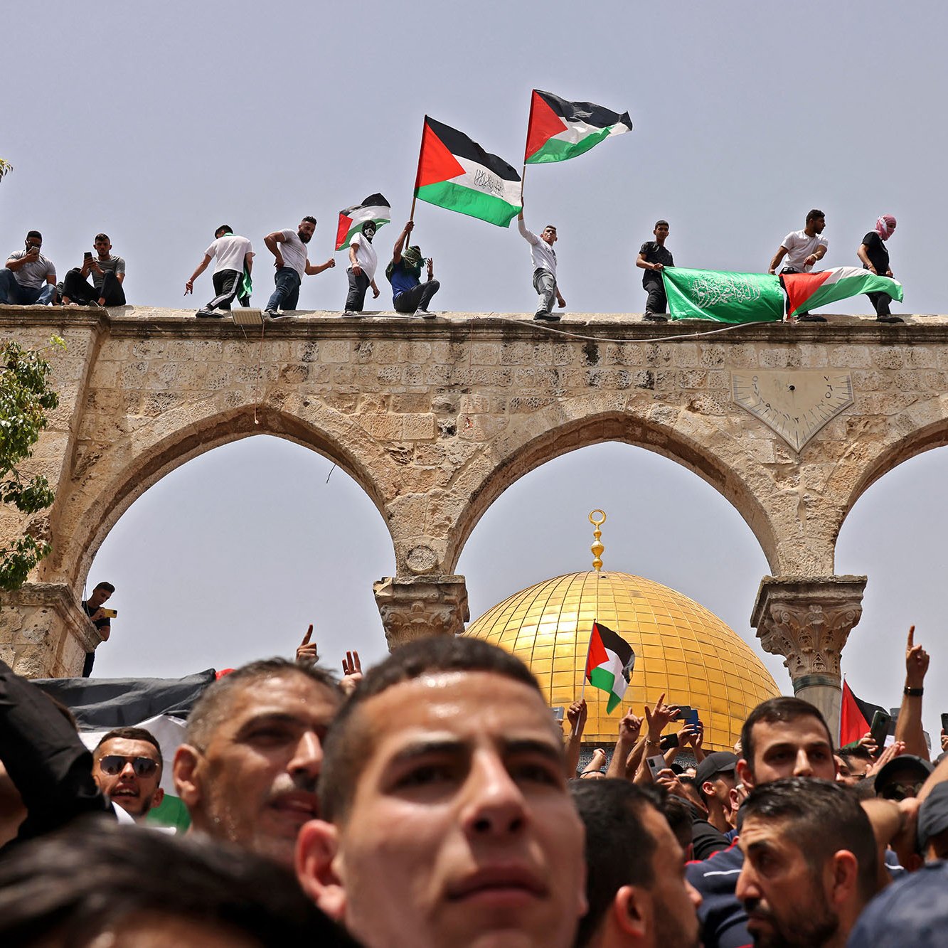 L'image montre une scène de rassemblement animé, avec un groupe de personnes portant des drapeaux palestiniens. Certaines personnes sont perchées sur un arc, tandis que la foule se trouve en dessous. En arrière-plan, on peut voir un dôme doré, probablement le Dôme du Rocher, un site emblématique de Jérusalem. L'atmosphère semble être celle d'une manifestation ou d'une célébration, avec une forte présence de symboles nationaux.