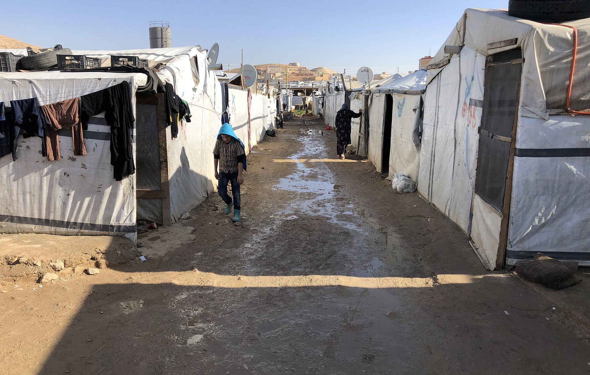 The image depicts a narrow path between rows of temporary tents or makeshift shelters, likely in a refugee or displaced persons camp. The tents are primarily white and appear to be made from tarpaulin or similar materials. There are clothes hanging outside some of the tents, suggesting attempts to dry them. The ground is muddy, indicating recent rain, and the surrounding area appears arid with few visible structures beyond the camp. The scene conveys a sense of modest living conditions and the challenges faced by individuals in such environments.