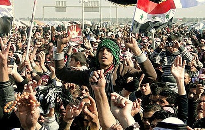 L'image montre une grande foule rassemblée, probablement lors d'une manifestation ou d'un événement public. Les personnes levées les bras, brandissant des drapeaux, affichent des expressions d'énergie et d'engagement. Certains portent des vêtements traditionnels, et l'ambiance semble dynamique et fervente. Des pancartes ou des symboles visibles pourraient suggérer un message politique ou social. L'environnement semble être un espace ouvert, peut-être une place ou une route.