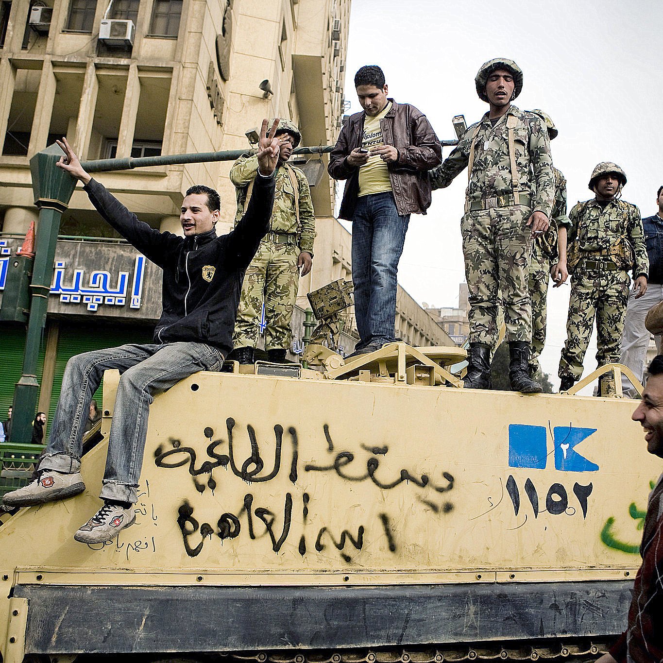 تظهر الصورة مجموعة من الأشخاص، بعضهم يرتدي زيًا عسكريًا، بينما الآخرون يرتدون ملابس عادية. يبدو أن هؤلاء الأشخاص متواجدون فوق مركبة عسكرية أو دبابه، حيث يعبر البعض عن مشاعرهم بإشارات وأيدي مرفوعة. يحاط بهم بعض المدنيين الذين يدفعون بحماس. الخلفية تحتوي على مباني وشوارع تشير إلى حضرية البيئة المحيطة. يمكن أن تعكس الصورة أجواء من التوتر أو الاحتجاجات.