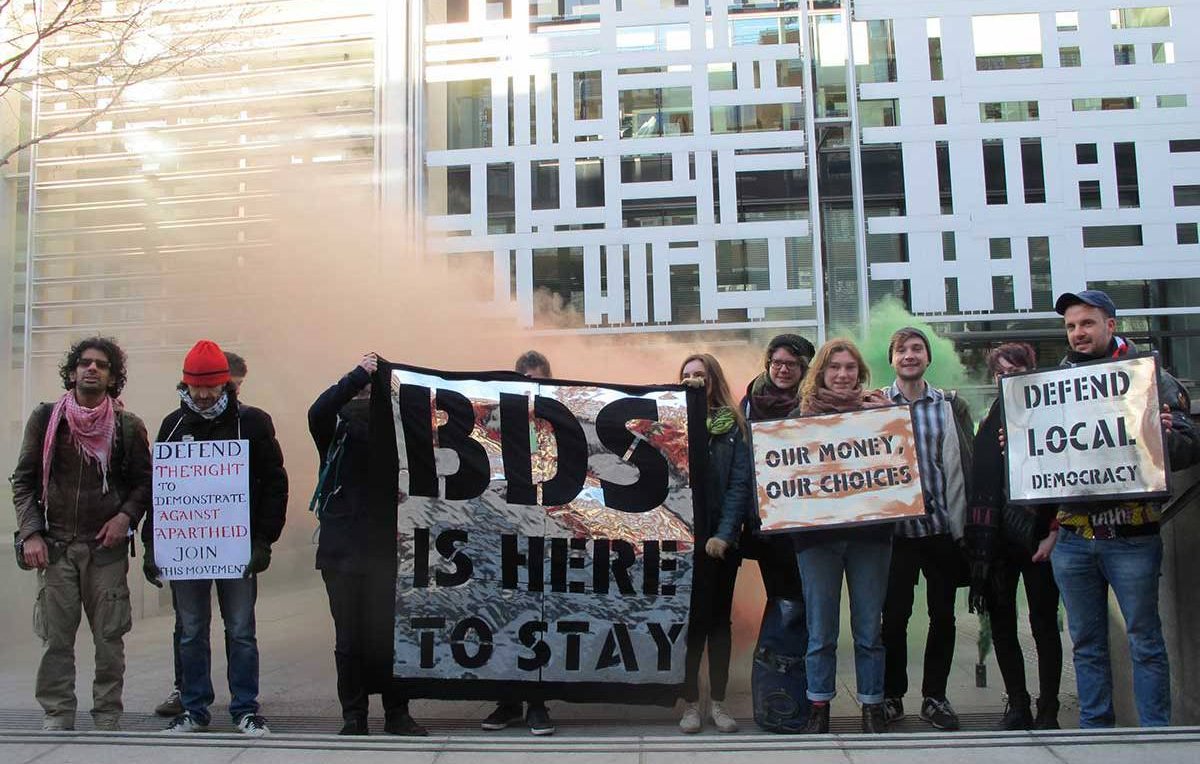 The image depicts a group of protesters standing outside a modern building, holding various signs. The signs convey messages related to support for the BDS movement, defending local democracy, and advocating for financial choices. The atmosphere seems charged, possibly with smoke or color in the background, emphasizing the protest's fervor. The group consists of diverse individuals, some wearing casual attire, and they appear to be unified in their cause.