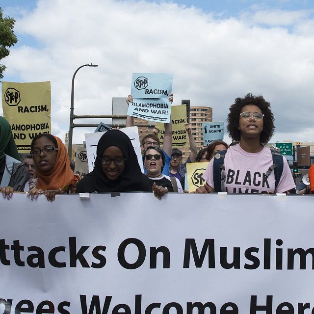 في الصورة، تظهر مجموعة من الأشخاص المشاركين في مظاهرة. يحمل المتظاهرون لافتات مكتوب عليها عبارات تدعو للتضامن مع المسلمين وترحب باللاجئين. يرتدي بعض النساء الحجاب، وتظهر تعابير الحماس والتأييد على وجوههم. السماء تبدو غائمة قليلاً، مما يضفي توتراً على الأجواء، وفي الخلفية يمكن رؤية مب buildings وشعارات تعبر عن رفض العنصرية والتمييز.