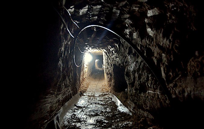 L'image montre un tunnel sombre creusé dans la terre. Les parois de la cavité sont en pierre, et le sol est inégal, probablement en terre battue. Une lumière blanche éclaire partiellement le tunnel, révélant son intérieur. Le fond du tunnel semble s'étendre à l'infini, créant une ambiance mystérieuse. Des câbles électriques sont visibles à certains endroits, indiquant qu'il pourrait y avoir des installations techniques à l'intérieur.