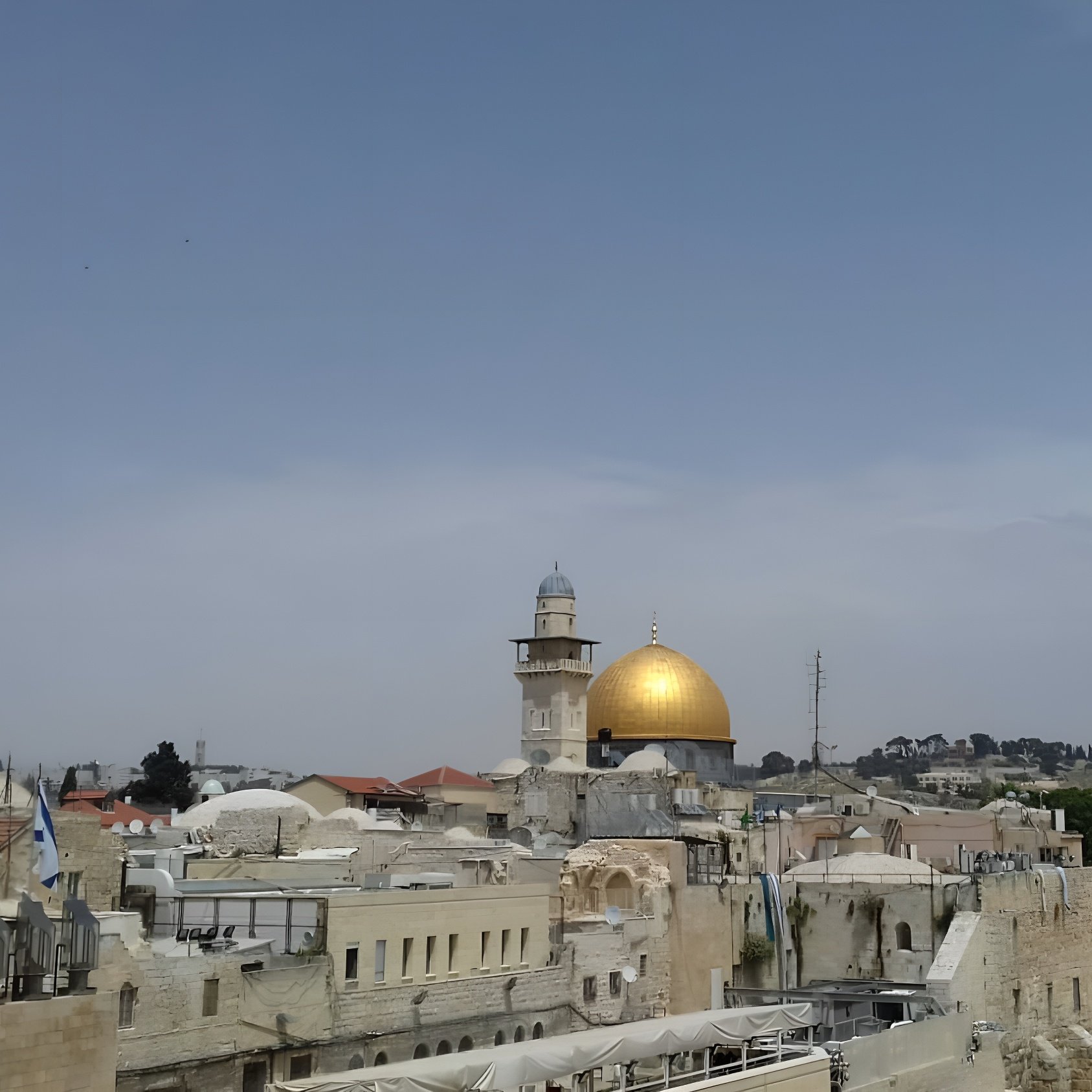 L'immagine mostra una vista panoramica di una città con una predominante cupola dorata, che è probabilmente il Dôme della Roccia, un noto sito religioso a Gerusalemme. Intorno ci sono edifici con tetti piatti e una torre, mentre il cielo appare sereno e leggermente nuvoloso. Gli alberi e le colline sullo sfondo aggiungono profondità alla scena.