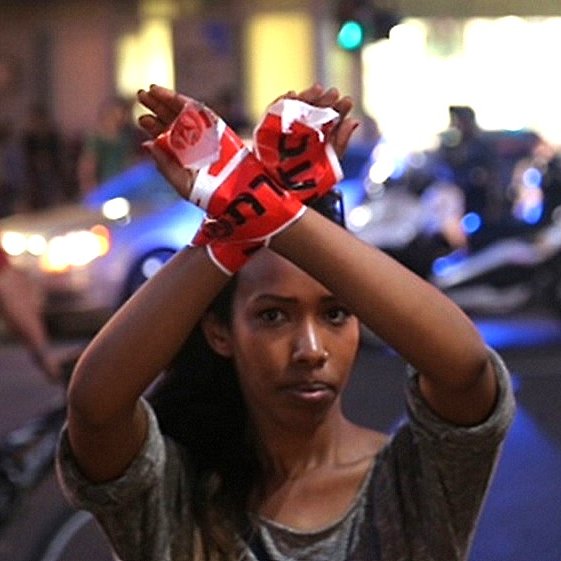 L'image montre une jeune femme qui se tient debout au milieu d'une rue, levant les bras avec des bandes de tissu rouge dans les mains, formant un symbole d'opposition ou de protestation. Elle semble déterminée et engagée, tandis que des véhicules et des lumières de police sont visibles en arrière-plan. Au fond, on peut apercevoir d'autres personnes, ajoutant un contexte urbain à la scène. L'atmosphère dégage un sentiment de mobilisation.