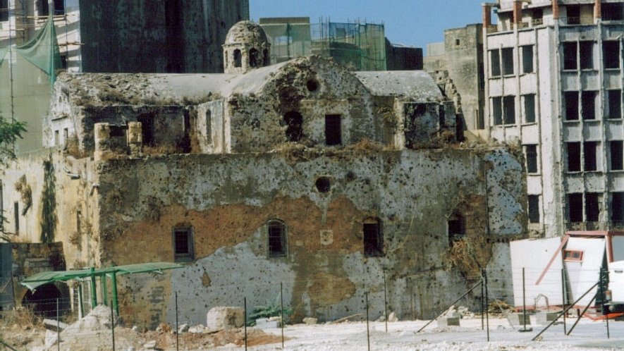 L'image montre des ruines d'un bâtiment ancien, probablement une église, dont la structure est en pierre et partiellement en décomposition. La façade est endommagée et recouverte de végétation, tandis qu'en arrière-plan, on peut voir des immeubles modernes en construction ou en rénovation. Le contraste entre l'ancien et le moderne donne une atmosphère de dégradation et de changement architectural. La zone semble être en chantier, avec des barrières et des échafaudages visibles.