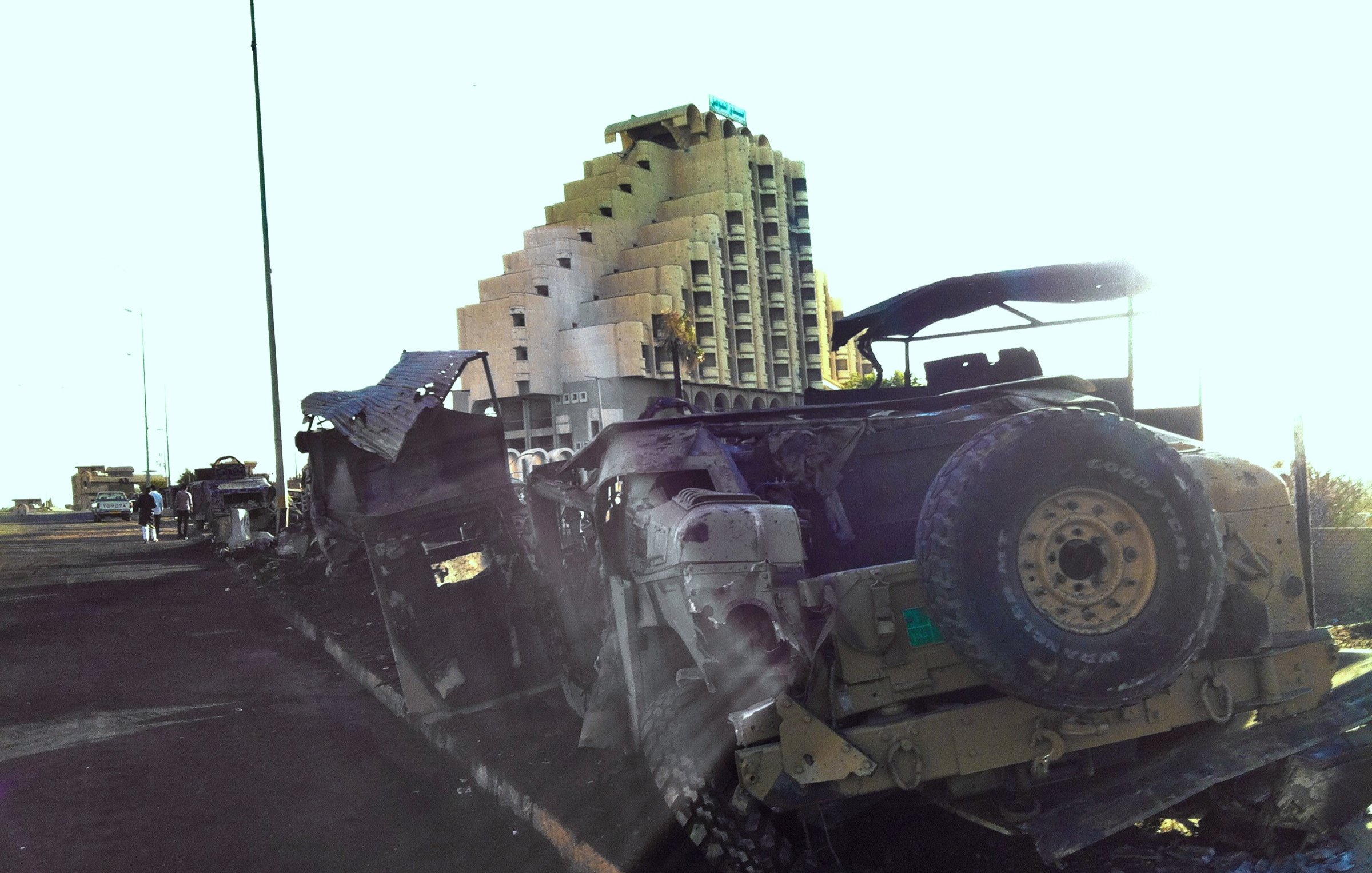 The image depicts a scene with damaged military vehicles, likely military trucks, that appear to be abandoned or destroyed along a road. In the background, there is a large, partially constructed building with a distinctive pyramid-like shape. The setting suggests a location that has experienced conflict or destruction, and the lighting gives the scene a bright, almost surreal quality. There are a few individuals visible in the background, indicating some activity in the area. Overall, the image conveys a sense of desolation and the remnants of past turmoil.