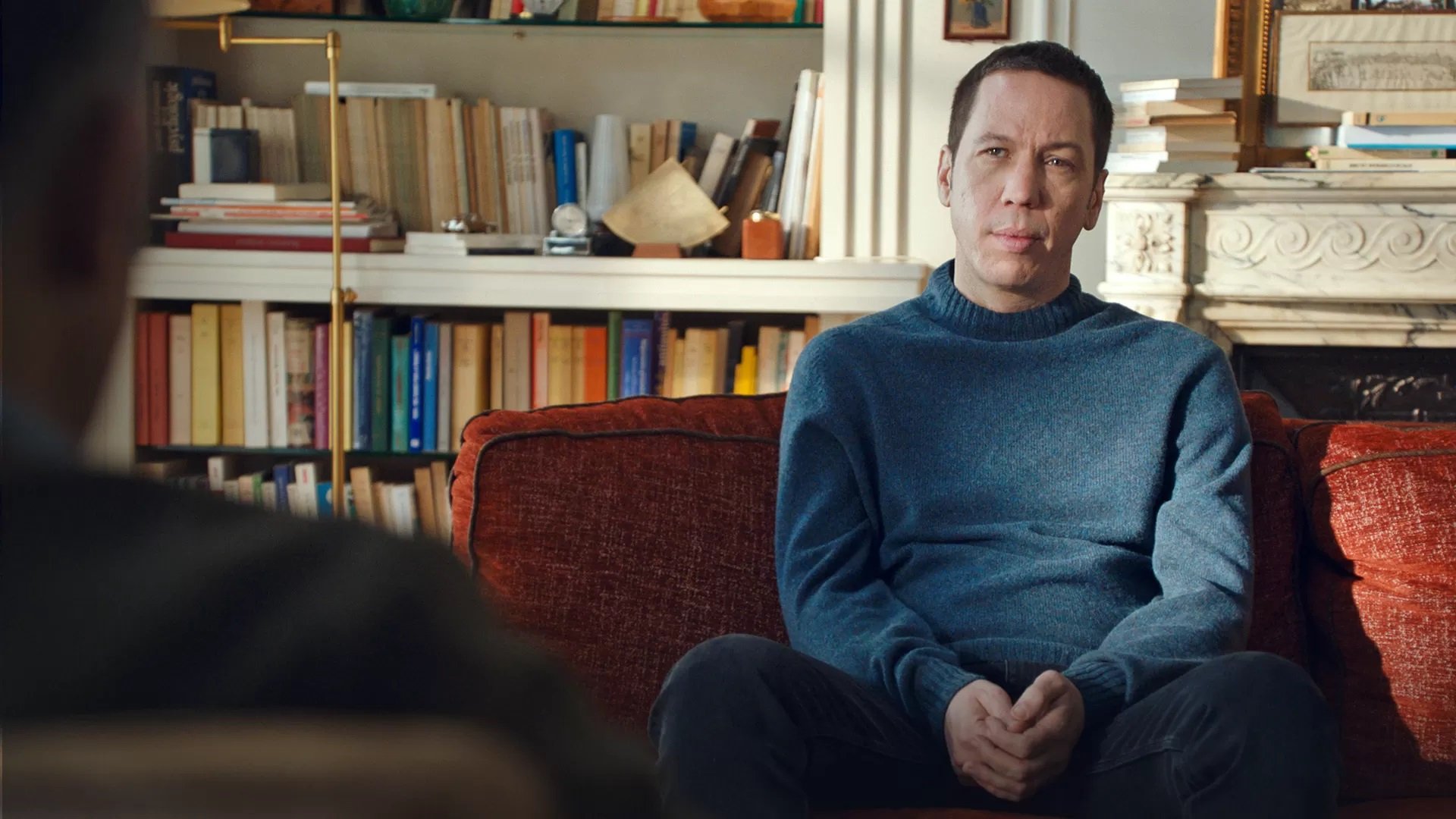 L'image montre un homme assis sur un canapé orange. Il porte un pull bleu et a une expression réfléchie. En arrière-plan, on peut voir une bibliothèque remplie de livres et quelques objets décoratifs. La lumière naturelle entre par une fenêtre, créant une ambiance chaleureuse et confortable dans la pièce. L'homme semble être en train d'écouter ou de participer à une conversation.