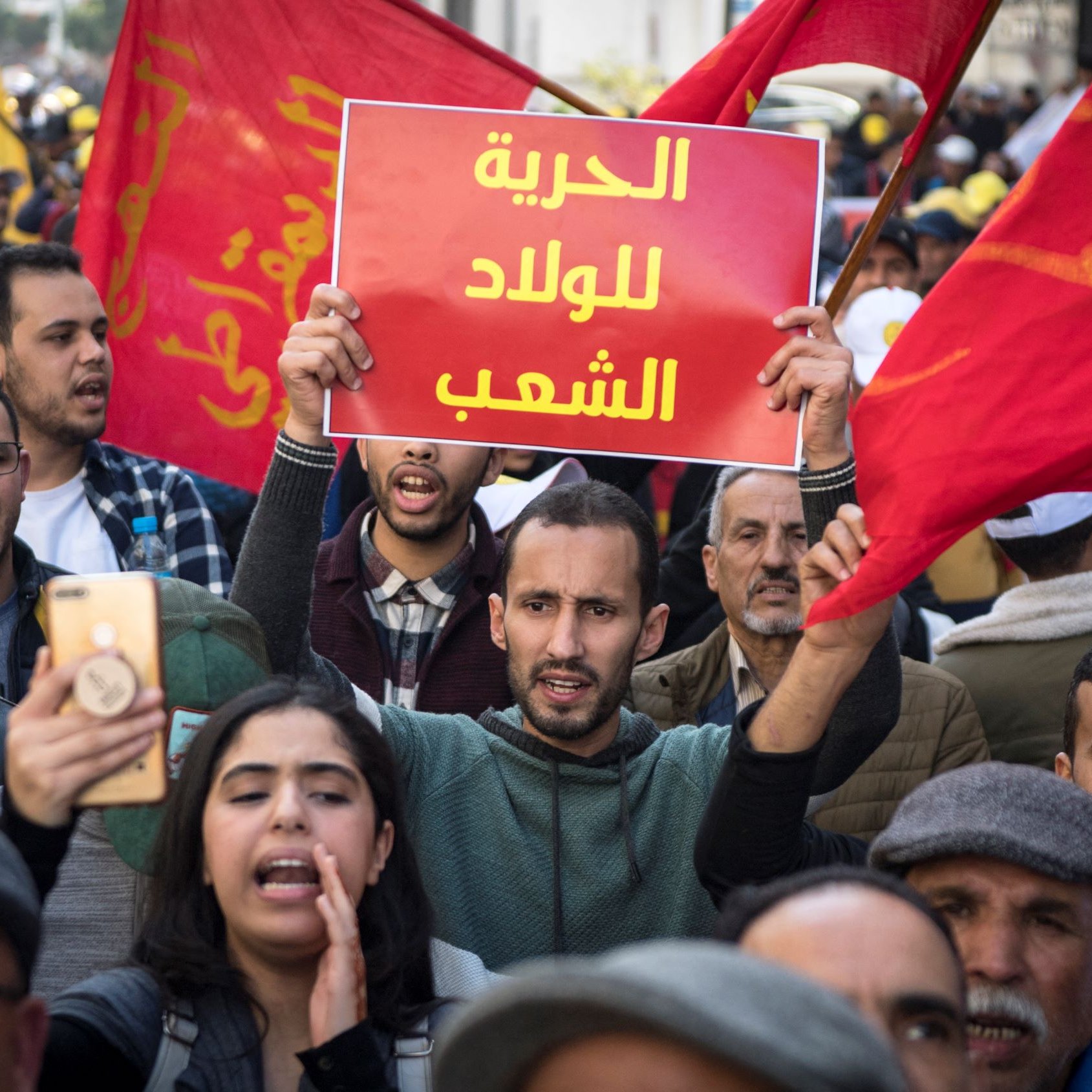 تظهر الصورة مجموعة من الأشخاص في مسيرة احتجاجية. يحمل أحد المتظاهرين لافتة مكتوب عليها "الحرية للولد الشعب"، بينما يبدو أن الحشود من حوله ترفع أعلامًا حمراء. التعبيرات في وجوه المتظاهرين تتراوح بين الحماس والعزيمة، مما يدل على روح التضامن والمشاركة في المطالبة بالحقوق. الجو العام يظهر فيه نشاط وحيوية، مما يشير إلى أهمية الرسالة التي يسعون لنقلها.