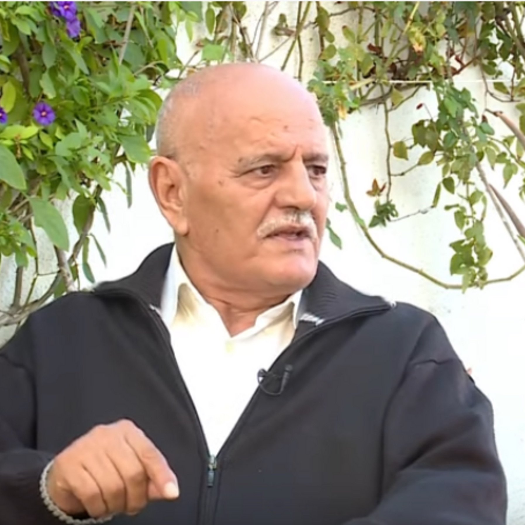 تظهر الصورة شخصًا في منتصف العمر يجلس في مكان خارجي، محاطًا بالنباتات الخضراء. يرتدي سترة داكنة ولديه لحية وشعر رمادي. يبدو وكأنه يتحدث أو يعبّر عن رأيه في موضوع ما، حيث يرفع إصبعه للتأكيد على نقطة سابقة. الخلفية تحتوي على نباتات وأزهار، مما يضفي جوًا طبيعيًا على المشهد.