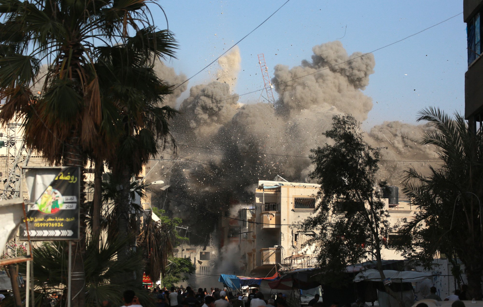 La imagen muestra una escena de destrucción y caos en una ciudad. Se puede ver una gran nube de polvo y escombros elevándose en el aire, lo que sugiere que ha ocurrido una explosión o un colapso de un edificio. En primer plano, hay un grupo de personas que parecen estar observando la situación, algunas de ellas con expresiones de sorpresa o preocupación. Alrededor, hay palmeras y estructuras urbanas que indican que se trata de un área urbana. El cielo es de un color azul claro, contrastando con el humo gris.