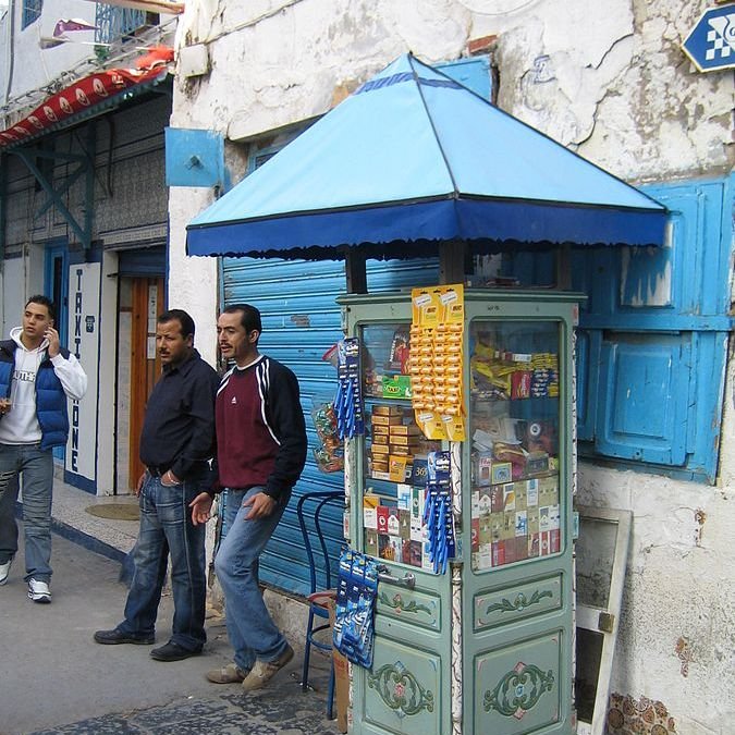 L'image montre un kiosque de vente situé sur un trottoir. Le kiosque, de style ancien, est peint en bleu et abrite divers articles comme des snacks, des boissons et des produits de consommation courante, visibles à travers les vitres. À côté du kiosque, il y a deux hommes. L'un porte un t-shirt, tandis que l'autre est habillé en noir. En arrière-plan, on peut voir des bâtiments aux murs usés et des volets bleus. L'atmosphère a l'air animée et typique d'un marché ou d'une rue commerçante.