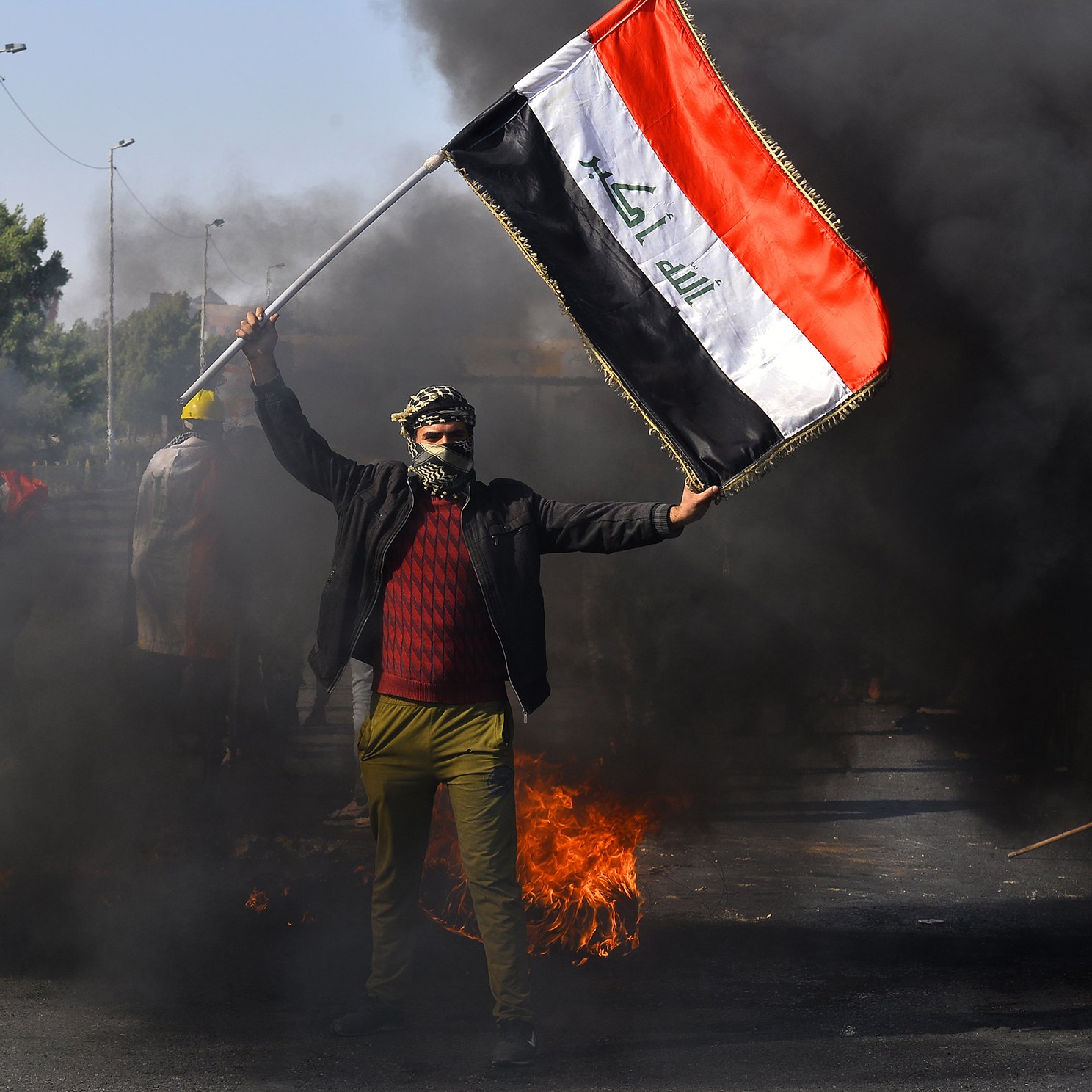The image depicts a person holding the Iraqi flag amidst a scene of protest. The individual is wearing a mask and has their face partially covered, standing in front of billowing black smoke and flames. The background suggests a chaotic environment, likely indicative of unrest or demonstrations. The focus on the flag emphasizes national identity in the context of the protest.