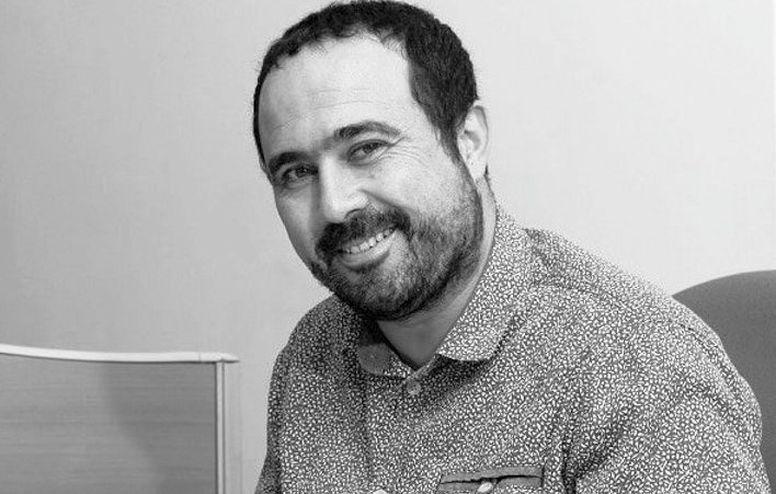 The image shows a man sitting and smiling. He has a short beard and is wearing a spotted button-up shirt. The background appears to be a plain wall and a piece of furniture, suggesting a professional or office setting. The overall tone of the image is friendly and approachable.