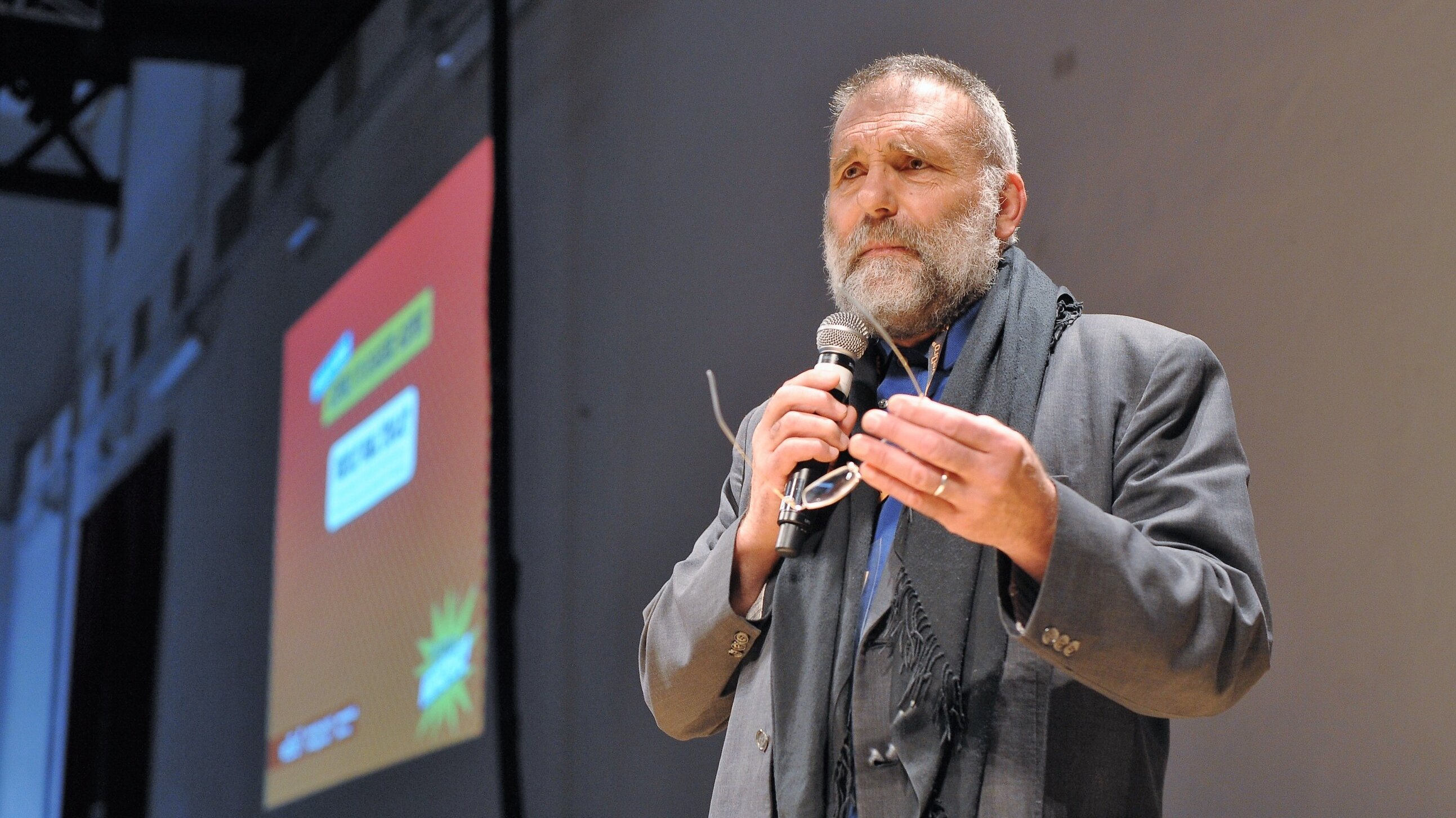 Nell'immagine si vede un uomo, probabilmente un relatore, che sta parlando al pubblico. Indossa un abito elegante e ha una sciarpa scura. Tiene un microfono in mano e sembra essere coinvolto in un discorso. Sullo sfondo c'è un grande schermo con delle grafiche colorate, ma il contenuto di ciò che viene mostrato non è visibile. L'atmosfera dell'immagine sembra essere formale e interattiva.