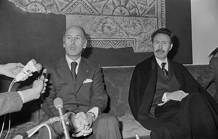 L'image montre deux hommes assis sur un canapé, apparemment lors d'une conférence de presse ou d'une rencontre. Ils semblent sérieux et attentifs, avec des microphones en face d'eux, indiquant qu'ils sont en train de répondre à des questions. L'un des hommes porte un costume sombre et a une expression réfléchie, tandis que l'autre, à sa droite, est vêtu d'une façon plus formelle, peut-être avec une écharpe ou un manteau. En arrière-plan, il y a un motif mural qui ajoute une certaine ambiance à la scène.