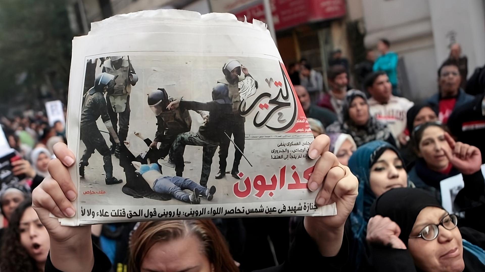 L'immagine mostra una manifestazione con un gruppo di persone unite in segno di protesta. In primo piano, una persona tiene in alto un giornale che riporta un'immagine significativa legata all'evento. La folla sembra esprimere emozioni forti, con alcune donne visibilmente agitatesi e altre che mostrano determinazione. Gli striscioni e le espressioni nei loro volti indicano il loro impegno per una causa importante. Il contesto generale evoca tensione e partecipazione attiva a una questione sociale.