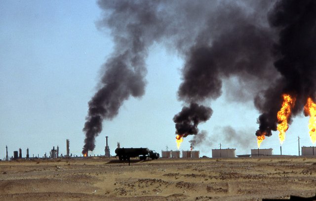 L'image montre un paysage désertique avec des installations industrielles en arrière-plan. On peut voir plusieurs cheminées émettant de la fumée noire dans le ciel, ainsi que des flammes s'élevant de réservoirs. Le sol est aride et ocre, accentuant le contraste avec la pollution visible. Cette scène évoque des activités liées à l'extraction ou au traitement du pétrole, suggérant des conséquences environnementales significatives.