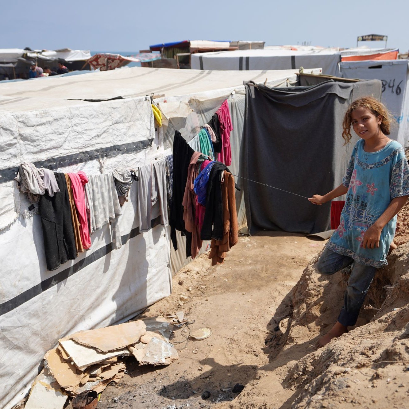 L'immagine ritrae un campo profughi improvvisato, con tende e strutture temporanee visibili in lontananza. In primo piano, c'è una giovane ragazza che sorride mentre si trova accanto a una tenda. I vestiti pendono da un filo tra le tende, e il terreno appare polveroso e irregolare. Sullo sfondo, il cielo è sereno e blu, creando un contrasto con la situazione circostante. La scena trasmette un senso di resilienza nonostante le difficoltà.