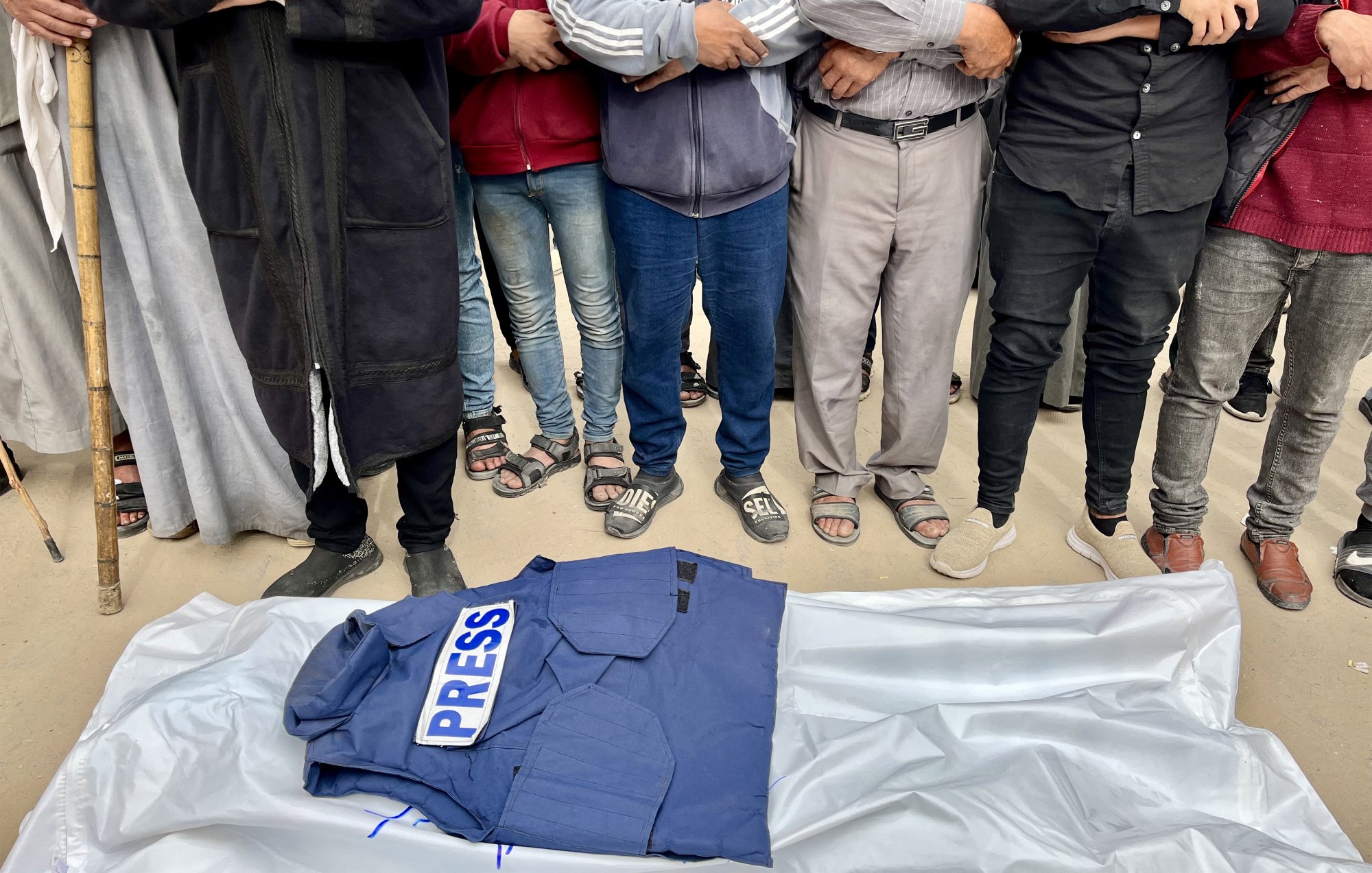 Des hommes entourent un gilet "PRESS" sur une bâche, en posture de recueillement.