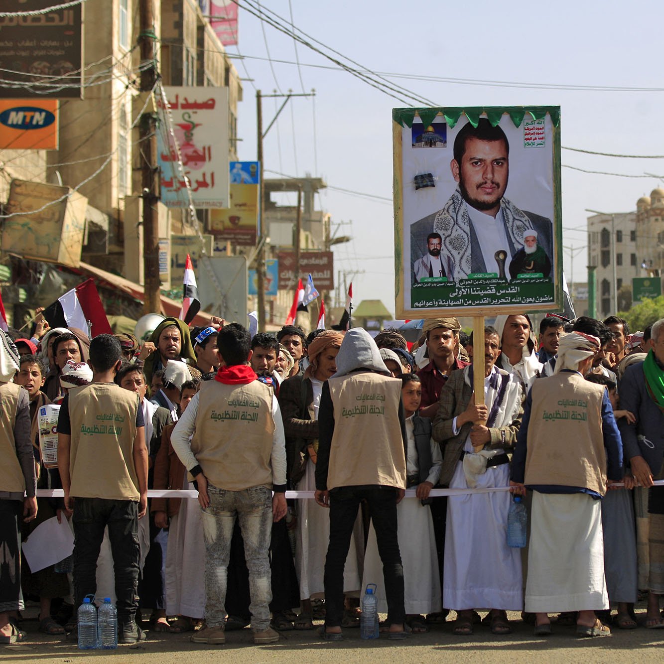 در این تصویر، گروهی از افراد در یک تظاهرات خیابانی حضور دارند. آنها لباس‌های مشابهی به تن دارند و تعدادی از آنها دارای چفیه هستند. در میان جمعیت، یک پوستر بزرگ از یک شخصیت سیاسی یا نظامی به نمایش گذاشته شده است که به وضوح جلب توجه می‌کند. پشت‌زمینه تصویر ساختمان‌هایی با نمای شهری دیده می‌شود و پرچم‌هایی از جمله پرچم یمن نیز در دست تظاهرکنندگان مشاهده می‌شود. جو کلی تصویر نشان‌دهنده یک تجمع سیاسی است.