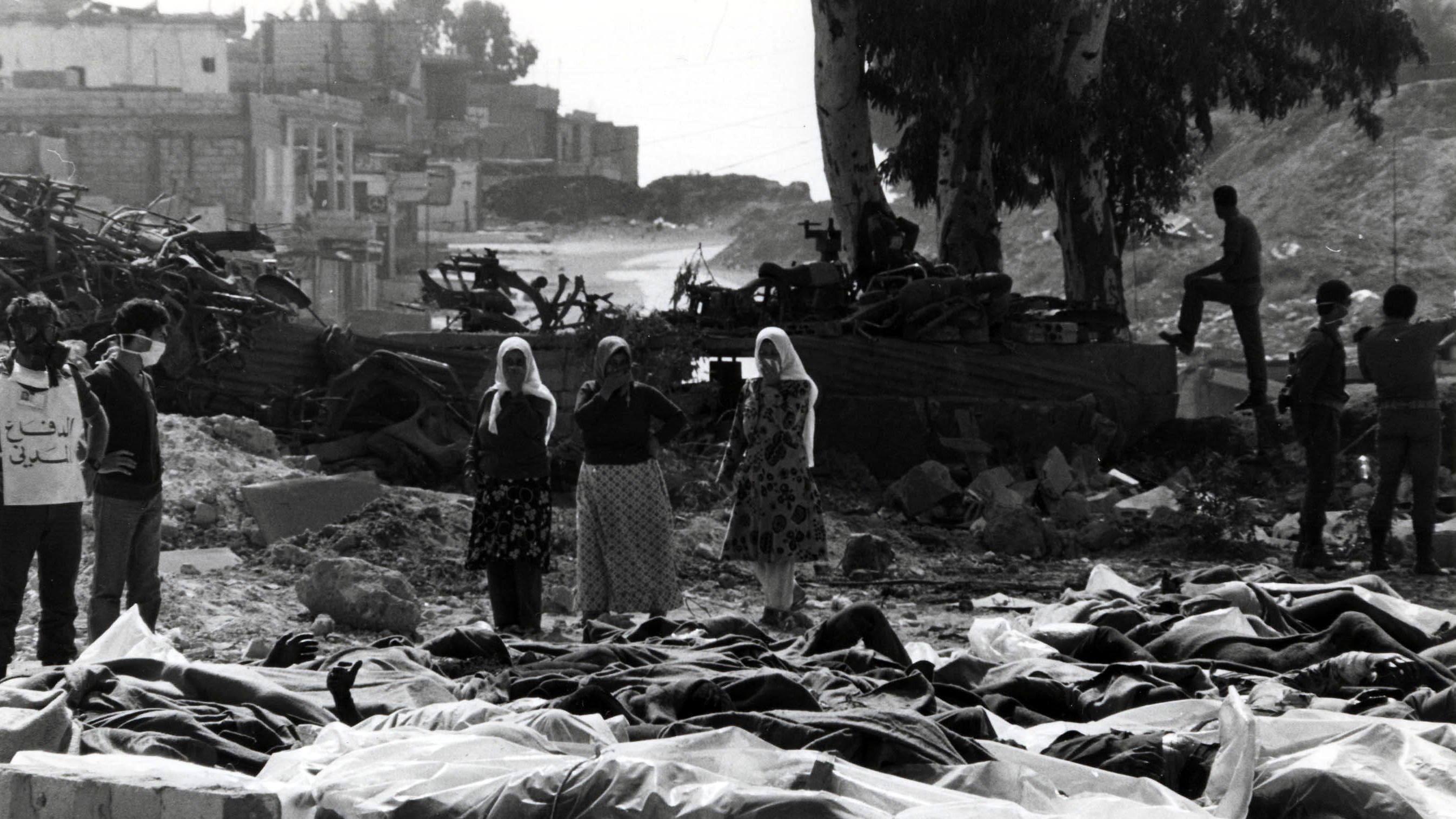 این تصویر نمایی غم‌انگیز و دل‌خراش از یک صحنهٔ بعد از بحران یا جنگ است. در جلوی تصویر، چندین جسد که با پارچه‌ای پوشیده شده‌اند، قرار دارند. در پس‌زمینه، گروهی از افراد حاضرند که به این صحنه نگاه می‌کنند، برخی از آنها اقدام به پوشاندن دهان و بینی خود کرده‌اند. درختان و ساختمان‌های ویرانه در پشت زمینه به چشم می‌خورند، که حاکی از تخریب و ویرانی است. جو کلی تصویر شعاری از درد و مصیبت را منتقل می‌کند.