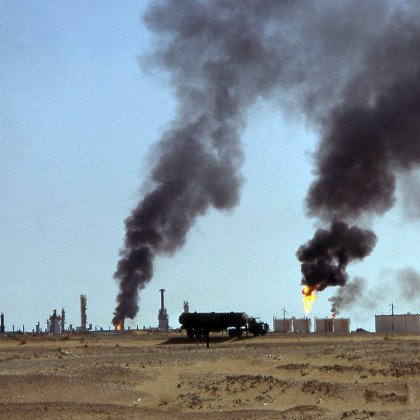 L'image montre un paysage désertique avec des installations industrielles en arrière-plan. On peut voir plusieurs cheminées émettant de la fumée noire dans le ciel, ainsi que des flammes s'élevant de réservoirs. Le sol est aride et ocre, accentuant le contraste avec la pollution visible. Cette scène évoque des activités liées à l'extraction ou au traitement du pétrole, suggérant des conséquences environnementales significatives.