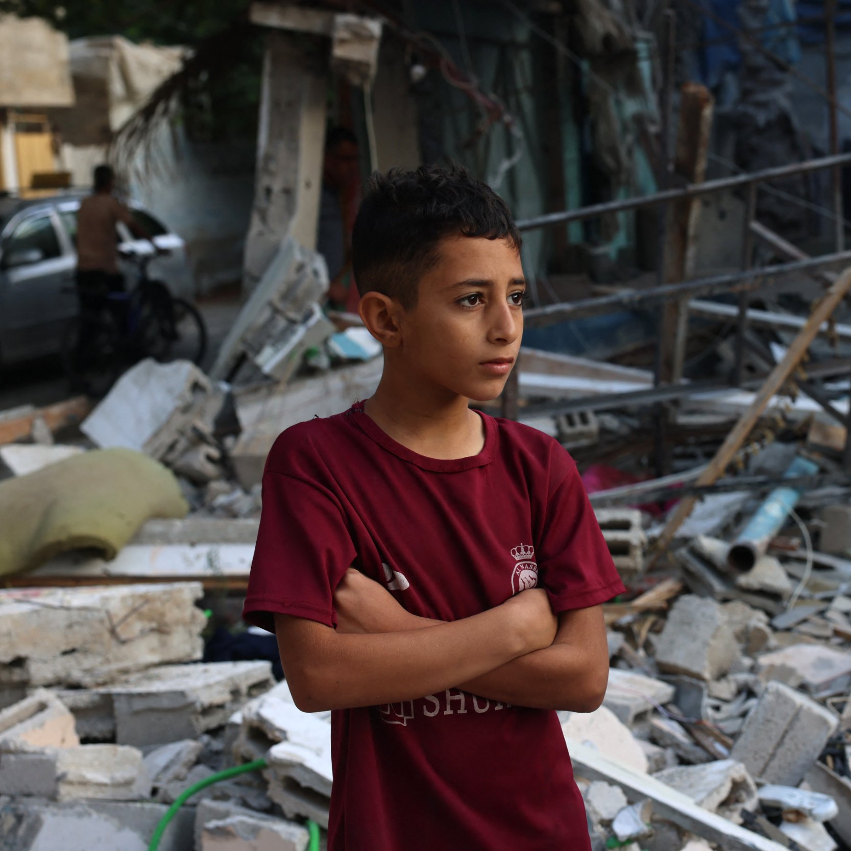 L'image montre un jeune garçon debout, les bras croisés, au milieu des débris d'un bâtiment effondré. Il semble pensif et préoccupé, exprimant une certaine gravité face à la destruction qui l'entoure. Les morceaux de béton et les matériaux épars suggèrent un environnement post-catastrophe, tandis que l'arrière-plan laisse entrevoir d'autres structures endommagées. L'atmosphère générale est sombre et témoigne d'une situation difficile.