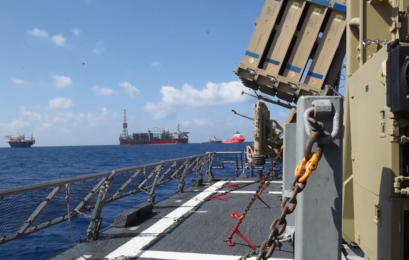 L'image montre un navire en mer, avec des structures de forage pétrolier visibles à l'horizon. On peut apercevoir un système de défense intégrée, probablement dédié à la surveillance ou à la protection, installé sur le pont du navire. Le ciel est clair avec quelques nuages, et la mer est calme, créant une atmosphère maritime paisible. Des chaînes et des équipements de sécurité sont également visibles sur le pont.